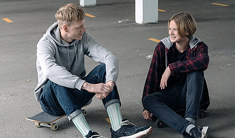 Zwei Jugendliche sitzend nebeneinander. Der Junge links sitzt auf einem Skateboard.