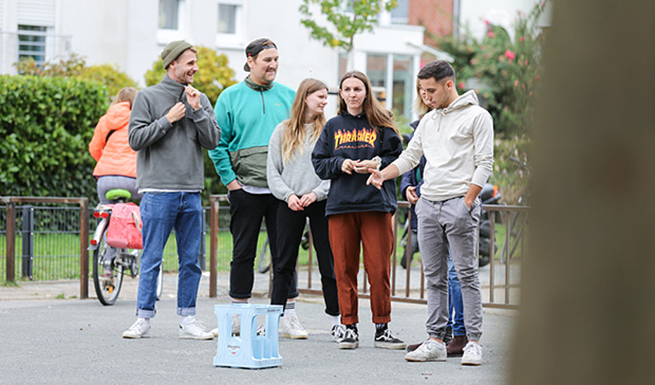 Fünf Jugendliche bei einem Gruppenspiel vor einem Flaschenträger der Straße stehend.