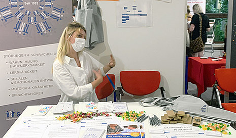 Eine Frau mit Mundschutz hinter einem Tisch mit Werbematerialien. Im Hintergrund ein Rollup von SeHT.grund 
