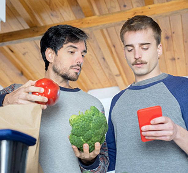 Zwei junge Männer nebeneinander. Der eine hält einen Brokkoli und eine Paprika. Der andere ein Handy, auf das beide schauen.